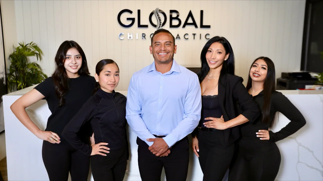 Five smiling staff members posing together in front of Global Chiropractic office sign